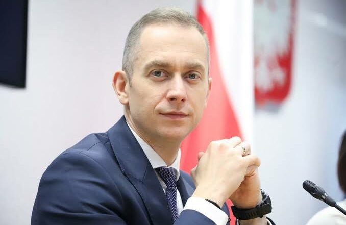 A man in a dark suit sits with clasped hands in front of a red and white flag and a microphone.