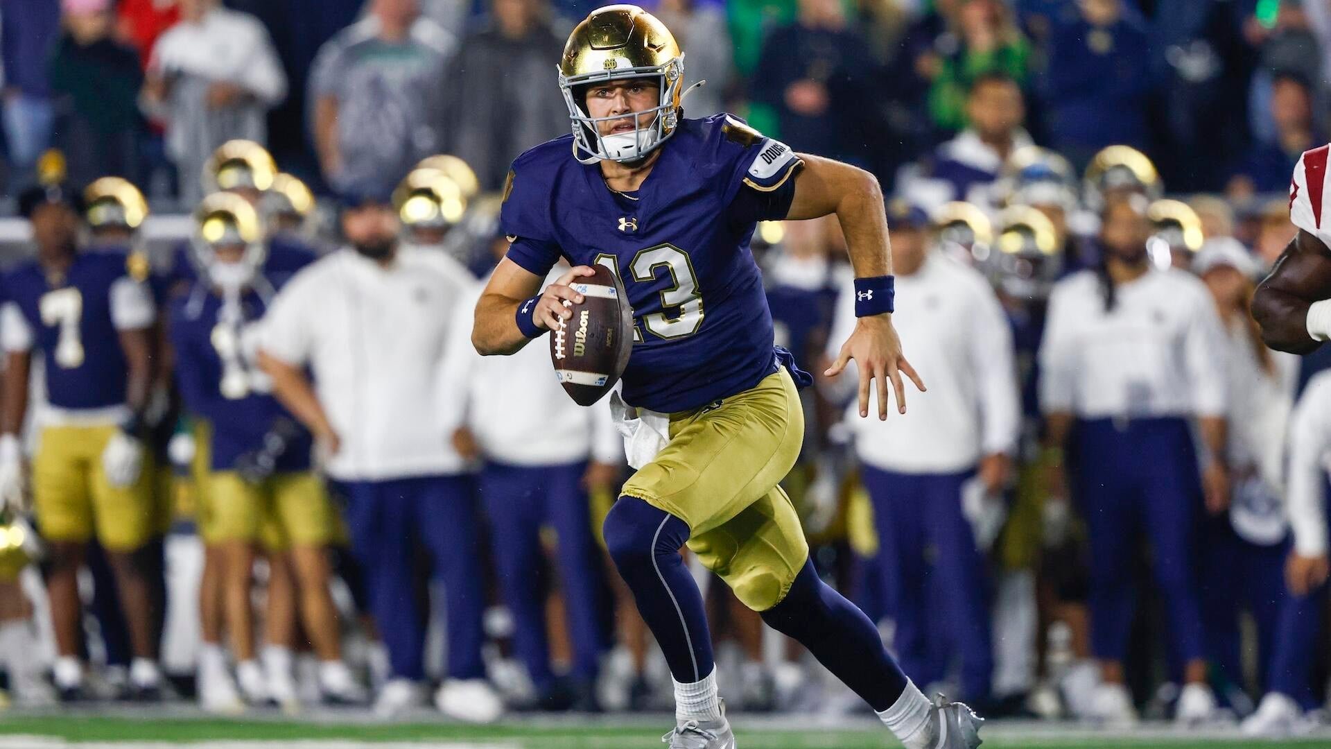 A football player wearing a navy blue and gold uniform with the number 13 runs with the ball tucked in his arm along the sideline.