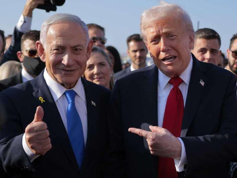 A bespectacled man in a navy blazer and blue shirt gestures with both hands while speaking in an office setting with shelves behind him.