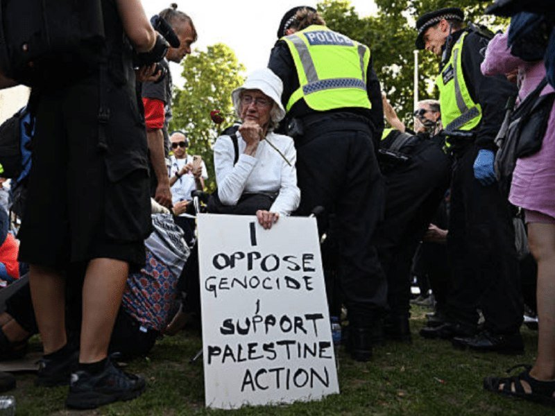 A seated protester holds a sign reading "I oppose genocide, support Palestine Action" while police officers in high-visibility vests stand nearby at an outdoor demonstration.