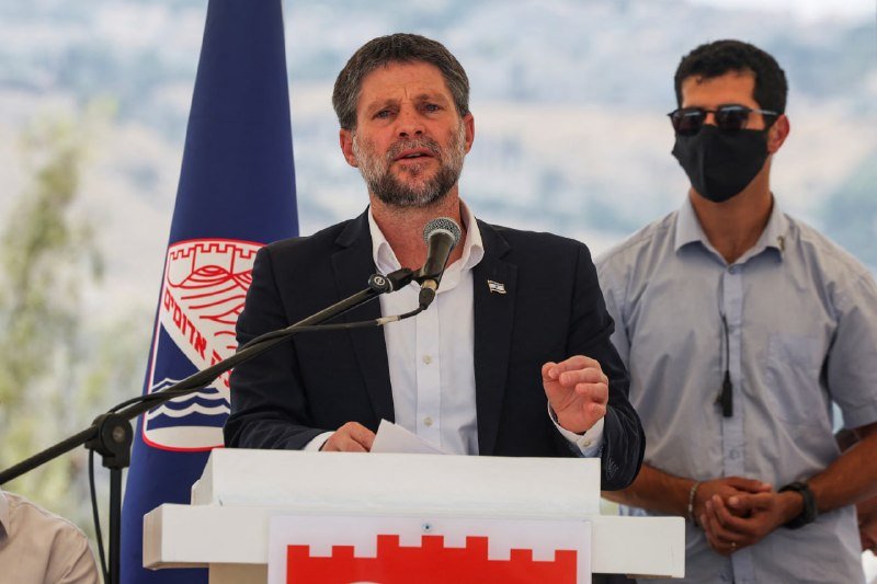 A bearded man in a dark suit speaks at a podium with microphones, gesturing with his hand, while a masked man in sunglasses and a light shirt stands behind him.