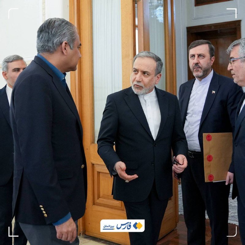 Five men in dark suits stand in a formal indoor setting, with one holding a folder bearing red seals.