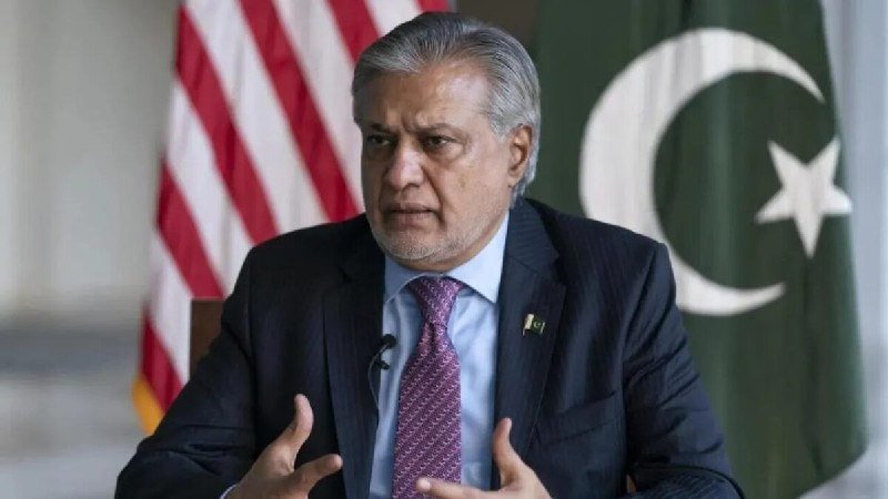 A gray-haired man in a dark suit and striped tie speaks while gesturing with his left hand during a meeting, with several national flags and microphones on the table.