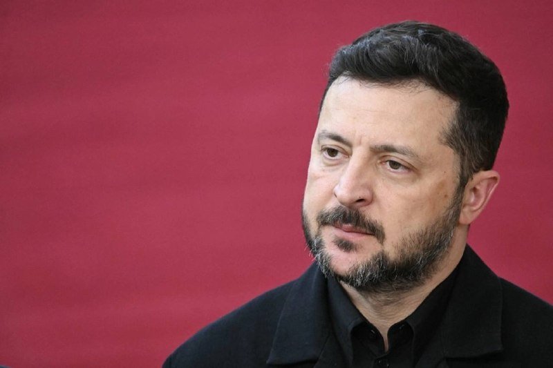 A bearded man with dark hair, wearing a black shirt and jacket, looks to the side against a solid magenta background.