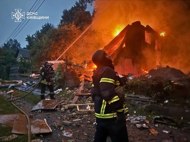 Several burnt-out, charred vehicles sit in a debris-strewn lot in front of a damaged brick building at night, with a "НАЦІОНАЛЬНА ПОЛІЦІЯ УКРАЇНИ" logo visible.