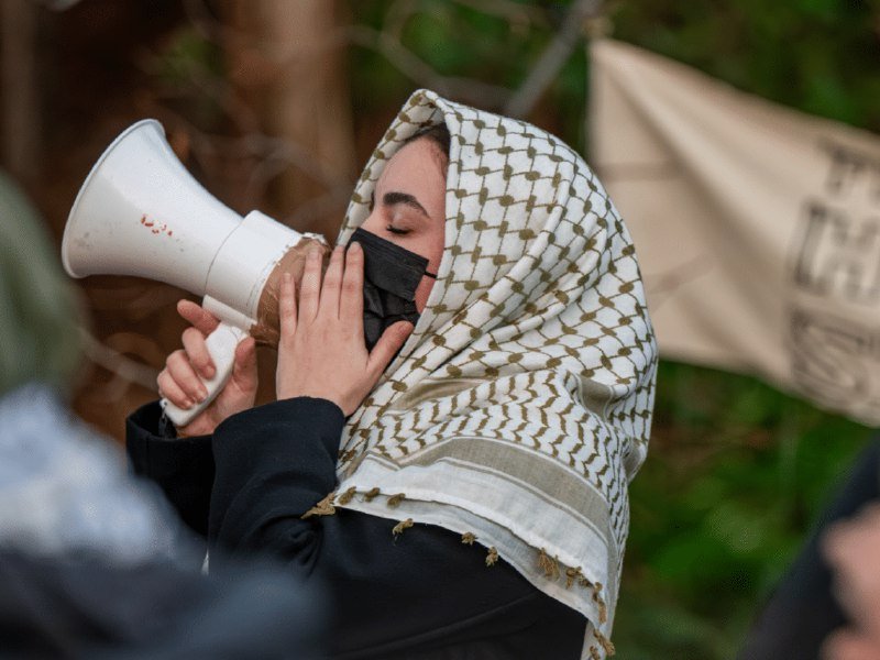 A masked person wearing a checkered keffiyeh speaks into a white megaphone during an outdoor demonstration, with a banner visible in the background.