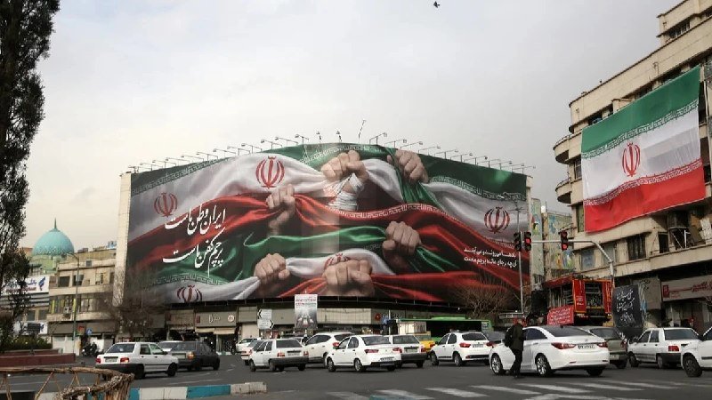 A large building displays a mural of fists gripping an Iranian flag, while a real Iranian flag hangs from an adjacent building along a busy street with cars and a pedestrian.