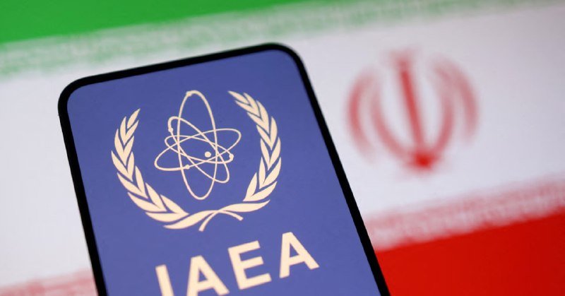 A smartphone displays the IAEA logo and text against a blurred background showing the Iranian flag.