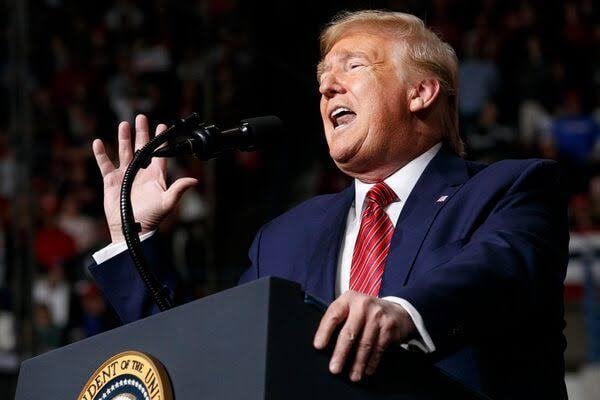 A man in a blue suit and red tie stands at a podium bearing the presidential seal, gesturing with one hand while speaking into a microphone before a crowd.