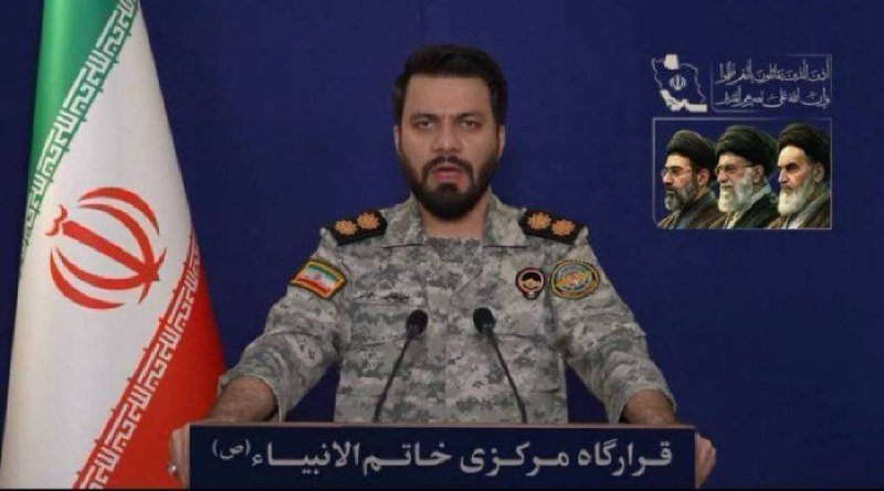 A bearded man in a camouflage military uniform speaks at a podium with microphones, with an Iranian flag and a portrait graphic visible behind him.