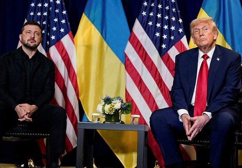 Two men in formal suits sit on chairs facing forward, with alternating American and Ukrainian flags displayed behind them and a small table holding flowers between them.