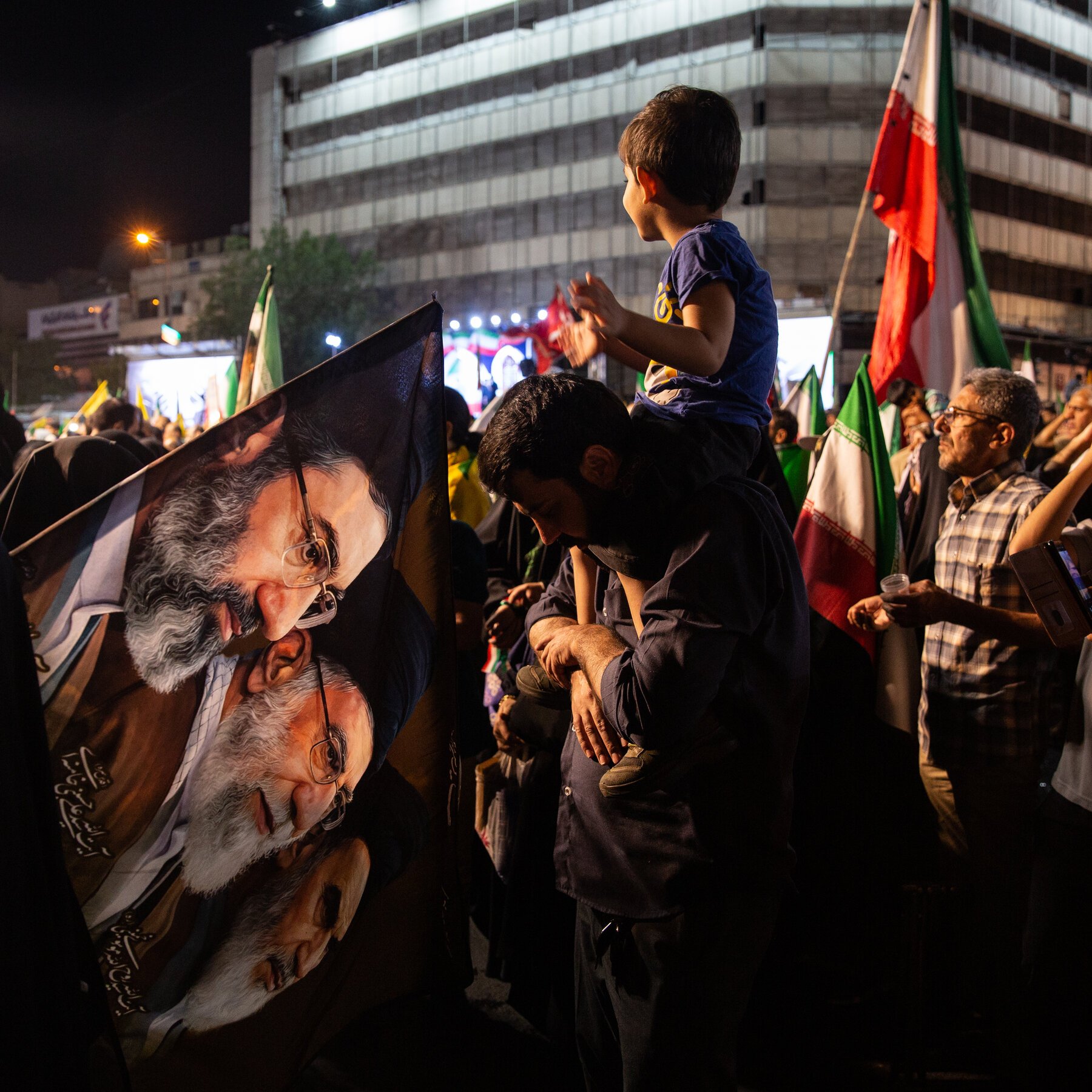 A man carries a child on his shoulders through a nighttime crowd holding flags and a banner bearing a portrait of a bearded man.