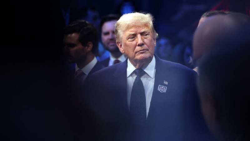 A split image shows two older men in dark suits; the left man has a white beard and wears a lapel pin, while the right man wears a striped tie near a microphone.