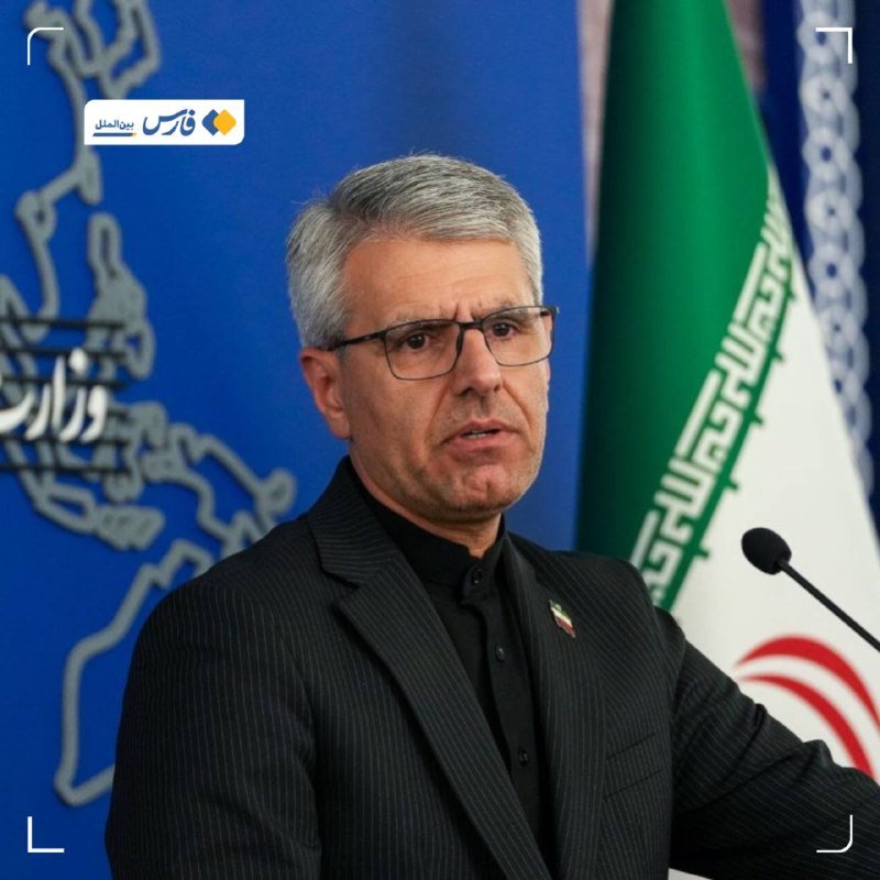 A gray-haired man wearing glasses and a dark pinstripe suit speaks at a microphone, with a map and an Iranian flag visible behind him.