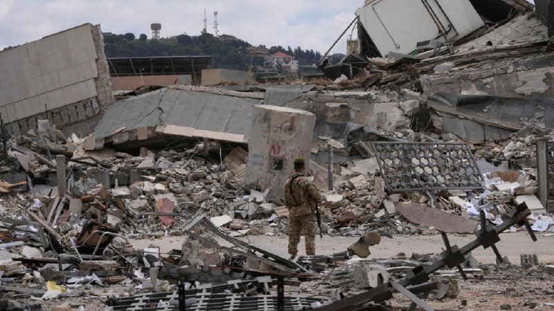 A soldier in camouflage stands amid extensive rubble and collapsed structures, with a graffiti-covered barrier and a communications tower visible on a hilltop in the background.