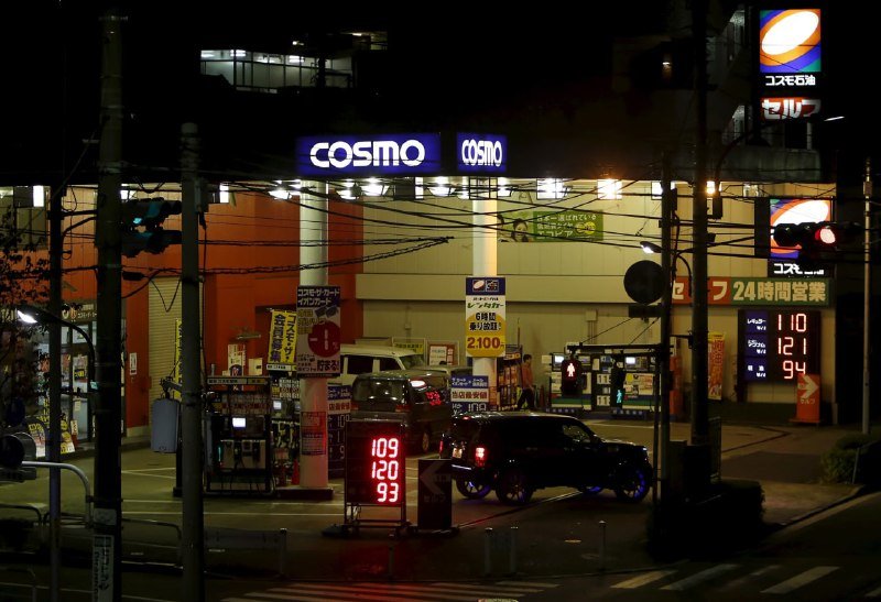 Nighttime street scene showing a COSMO gas station, illuminated digital price signs displaying fuel costs, and vehicles passing through an intersection.