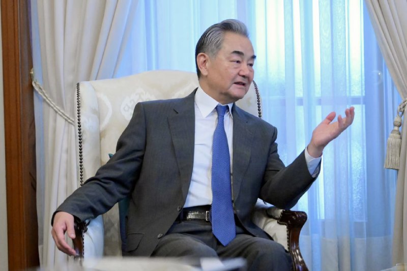 A gray-haired man in a dark suit speaks and gestures with his hand at a conference table featuring small flags, microphones, water bottles, and papers.