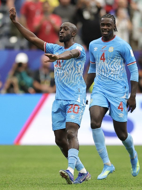 Two soccer players in light blue kits with numbers 20 and 4 celebrate on a green pitch, one with arms outstretched, while spectators and photographers watch from the blurred background.