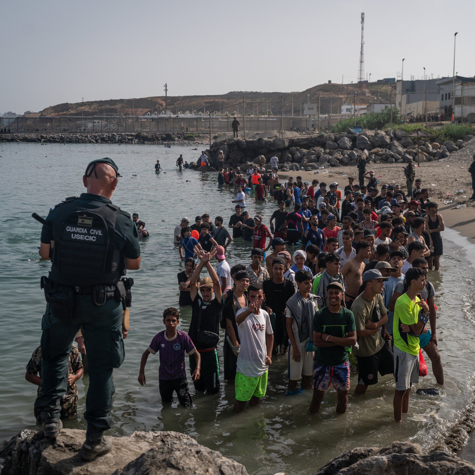 A Guardia Civil officer watches as a large crowd of people wade through shallow coastal water toward a rocky shoreline.