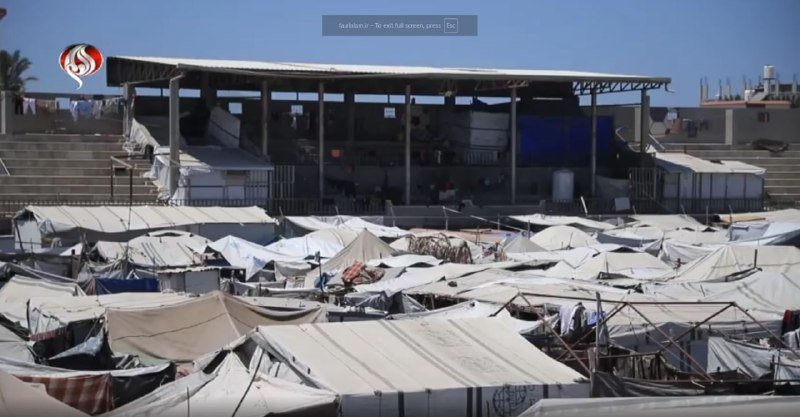 A sprawling encampment of densely packed white tents stretches beneath a large open-sided pavilion structure under a clear blue sky.