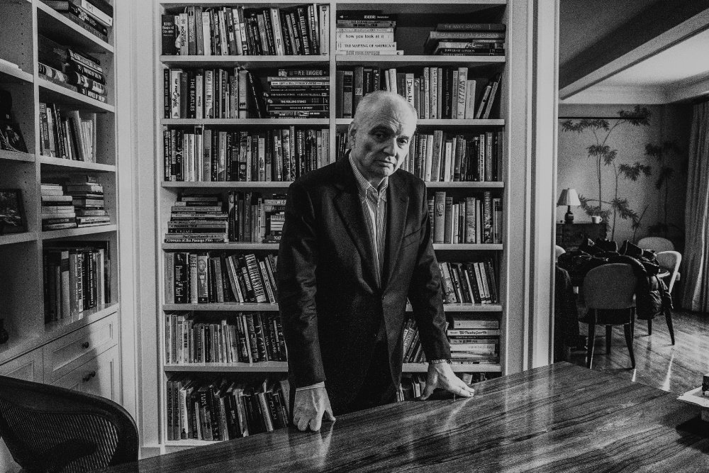 A black-and-white photo shows a gray-haired man in a dark blazer standing with both hands resting on a wooden table, backed by tall bookshelves filled with books.