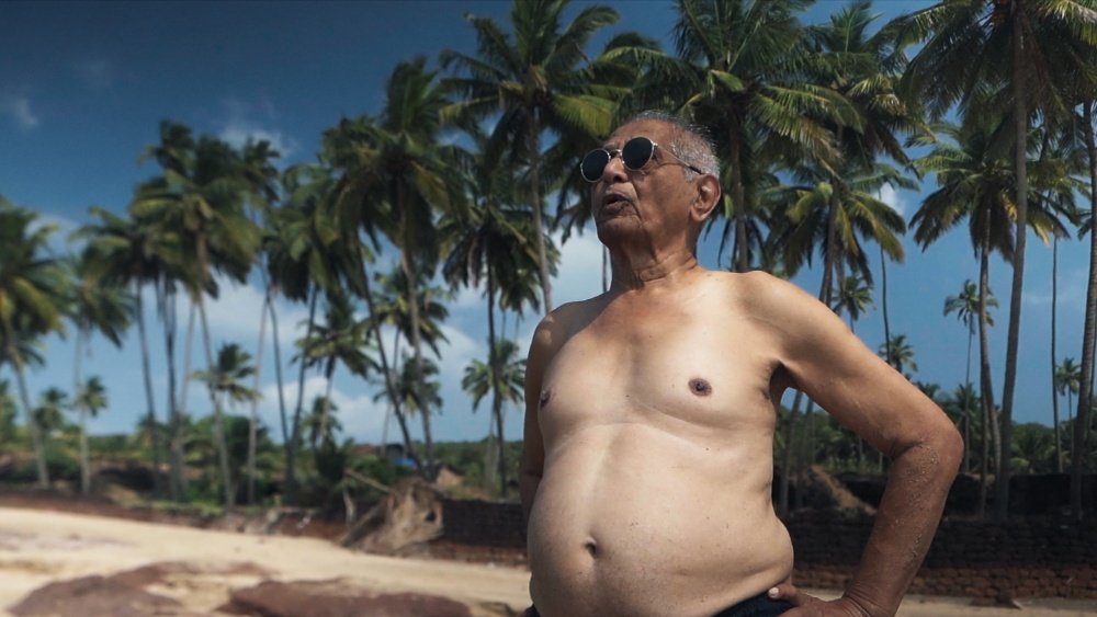 A shirtless older man wearing sunglasses stands with his hand on his hip against a backdrop of tall palm trees and a partly cloudy sky.