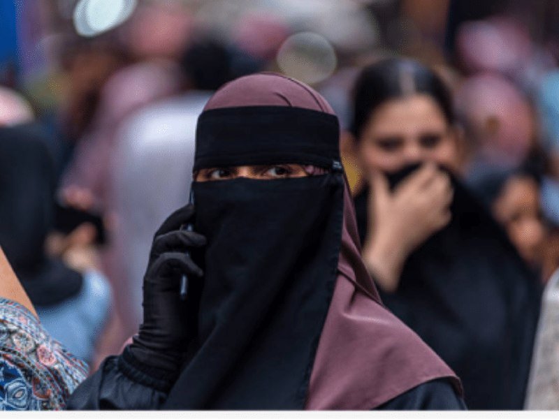 A woman wearing a black niqab and gloves holds a phone to her ear amid a blurred crowd.