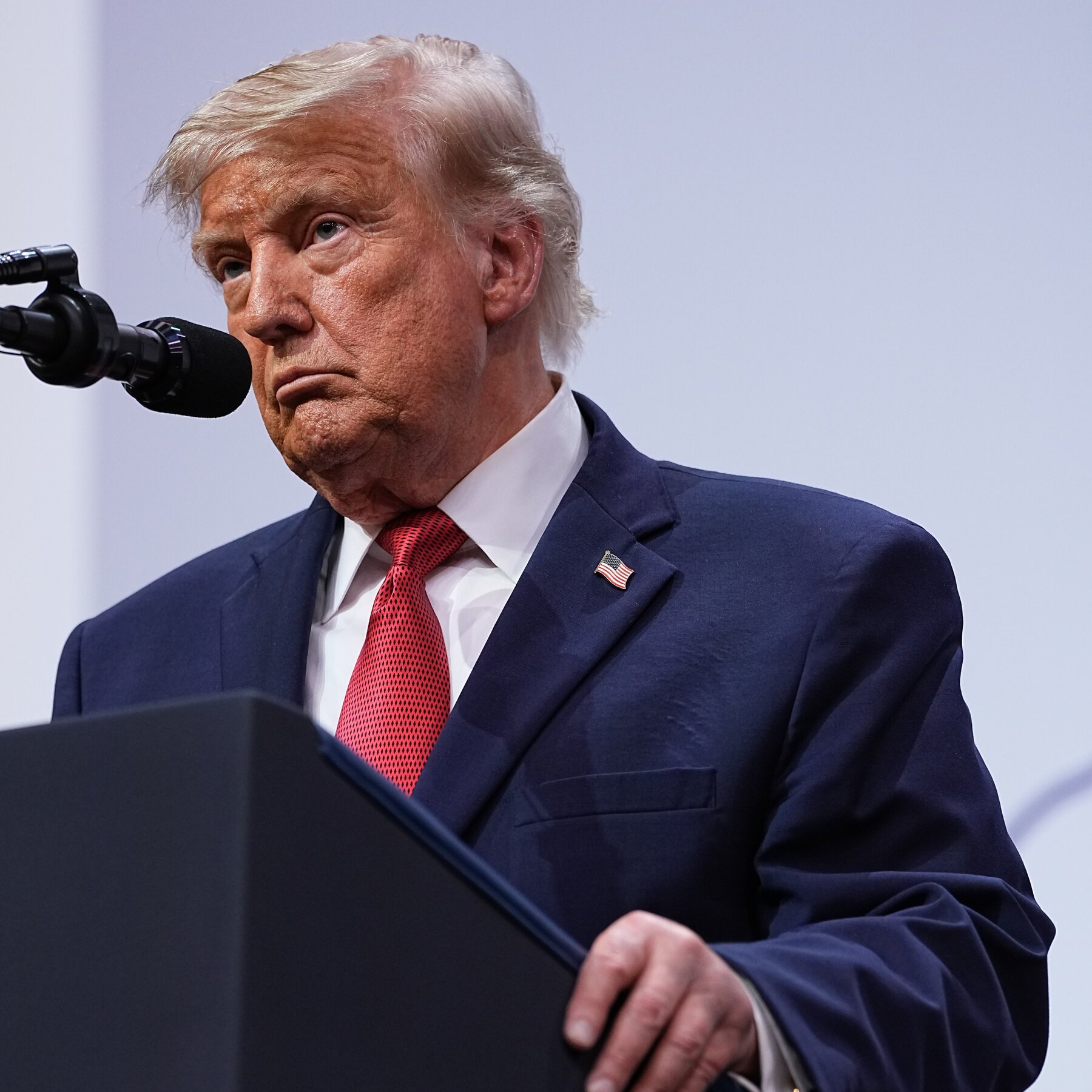 An older man with blond hair, wearing a navy suit, red tie, and American flag lapel pin, stands at a podium near a microphone.