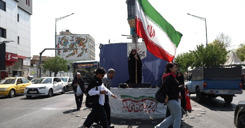 A woman waves a large Iranian flag on a decorated platform in the middle of a city street, while pedestrians walk past and cars travel along the road.
