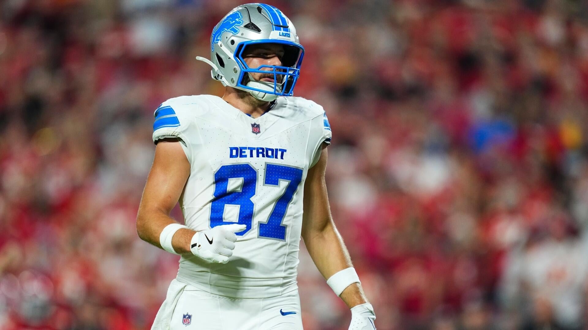 A Detroit Lions football player wearing jersey number 87 and a silver helmet stands on the field against a blurred crowd backdrop.