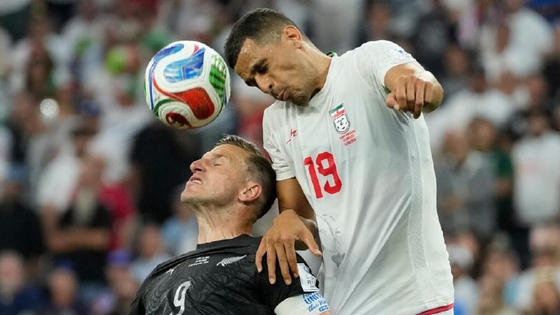 A goalkeeper in pink dives toward a ball near the goal line during a soccer match, with players in white and black jerseys watching and fans holding Iranian flags in the stands.