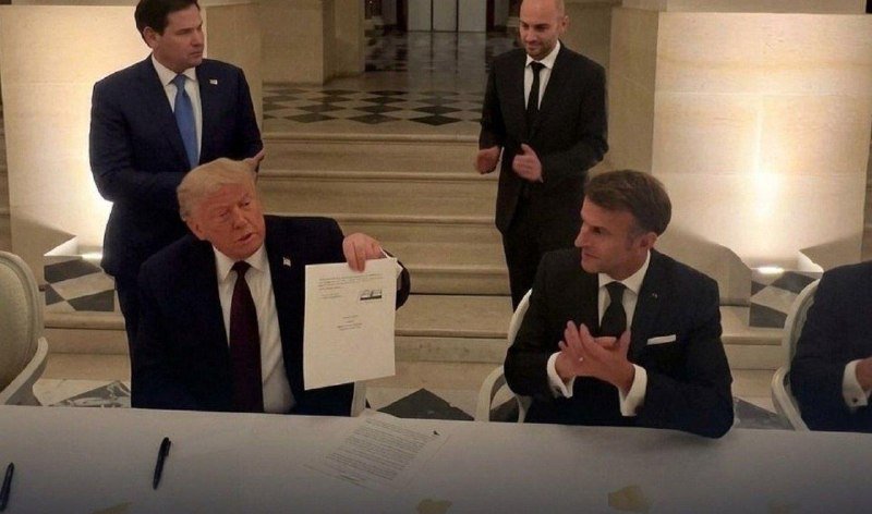 Two men in suits sit at a table as one holds up a document; two other men in suits stand behind them in a formal, marble-floored room.