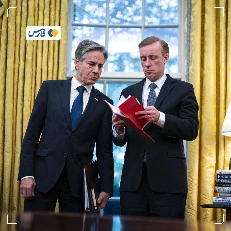 Two men in dark suits stand in a yellow-curtained room, one holding and reviewing a red folder while the other looks on, beside a window.