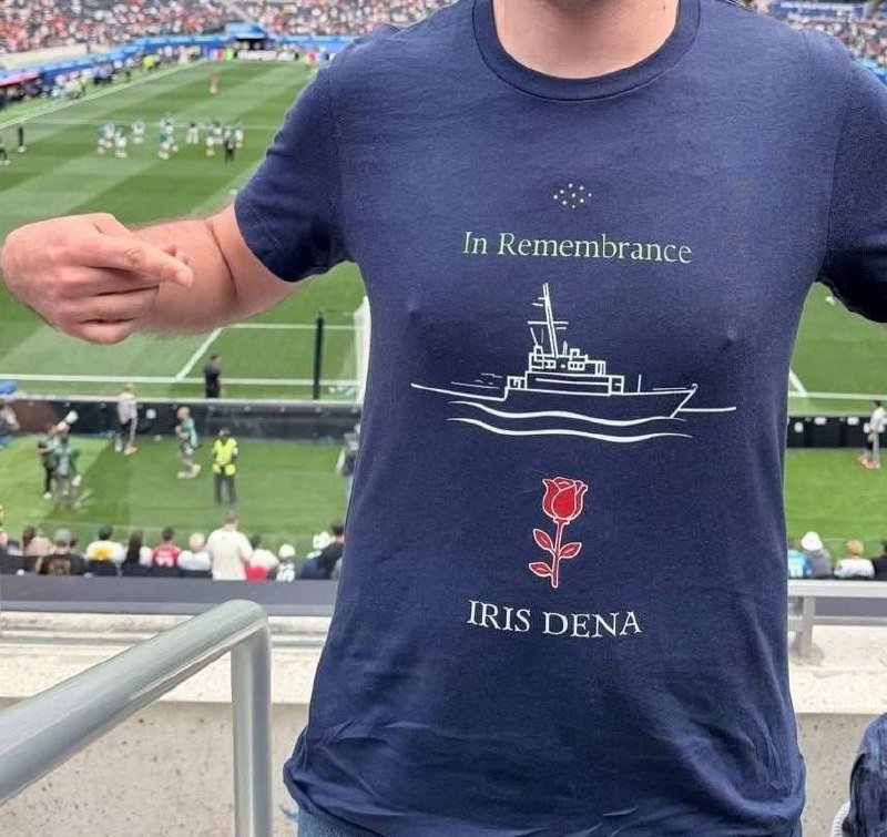 A person stands in a stadium wearing a navy "In Remembrance" t-shirt featuring a ship illustration, red rose, and "IRIS DENA" text, with a soccer match visible on the field below.