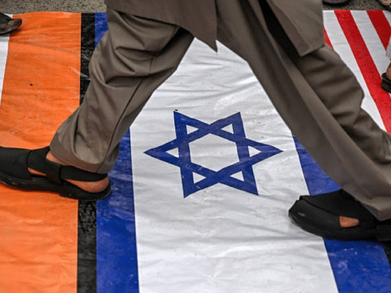 Four men in suits stand on a stage before American and other flags, with one man clapping and another walking forward.