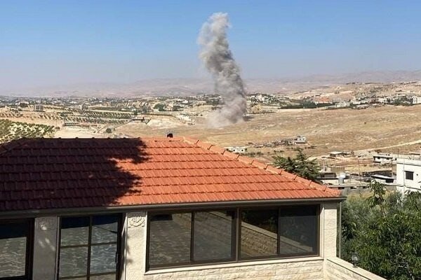 A plume of smoke rises over a sprawling Middle Eastern town viewed past the red-tiled roof of a nearby building.