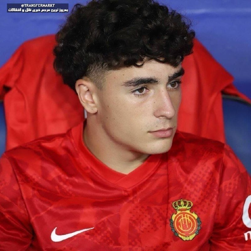 A young man with curly dark hair sits wearing a red athletic jacket featuring a Nike logo and a crowned crest emblem.