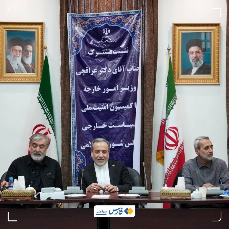 Three men sit at a conference table flanked by Iranian flags, with a Persian banner and two framed portraits of bearded clerics hanging behind them.