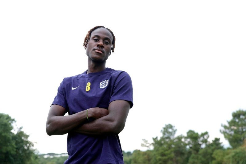 A young man with braided hair stands outdoors with arms crossed, wearing a dark blue soccer jersey featuring a Nike logo and a three-lions crest.