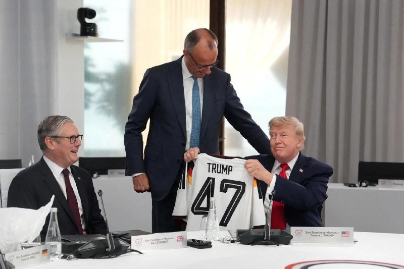 Three men in suits at a conference table; one seated holds up a white jersey reading "TRUMP 47" while two others look on smiling.
