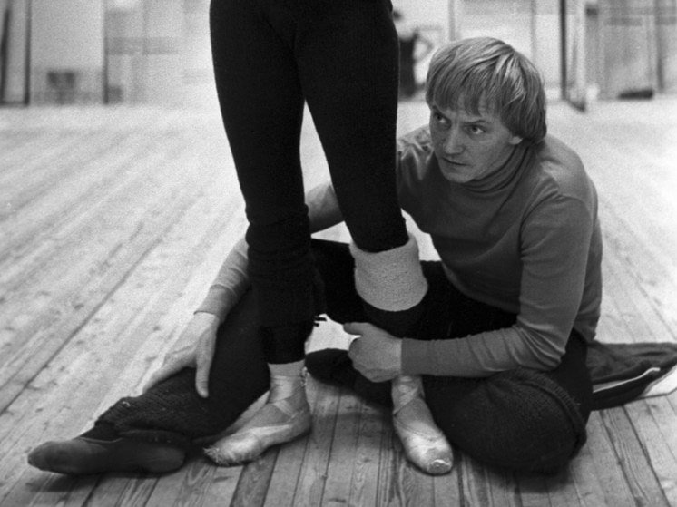 Black-and-white photo of a man in a turtleneck kneeling on a wooden floor, adjusting the ankle of a dancer in tights and pointe shoes during rehearsal.