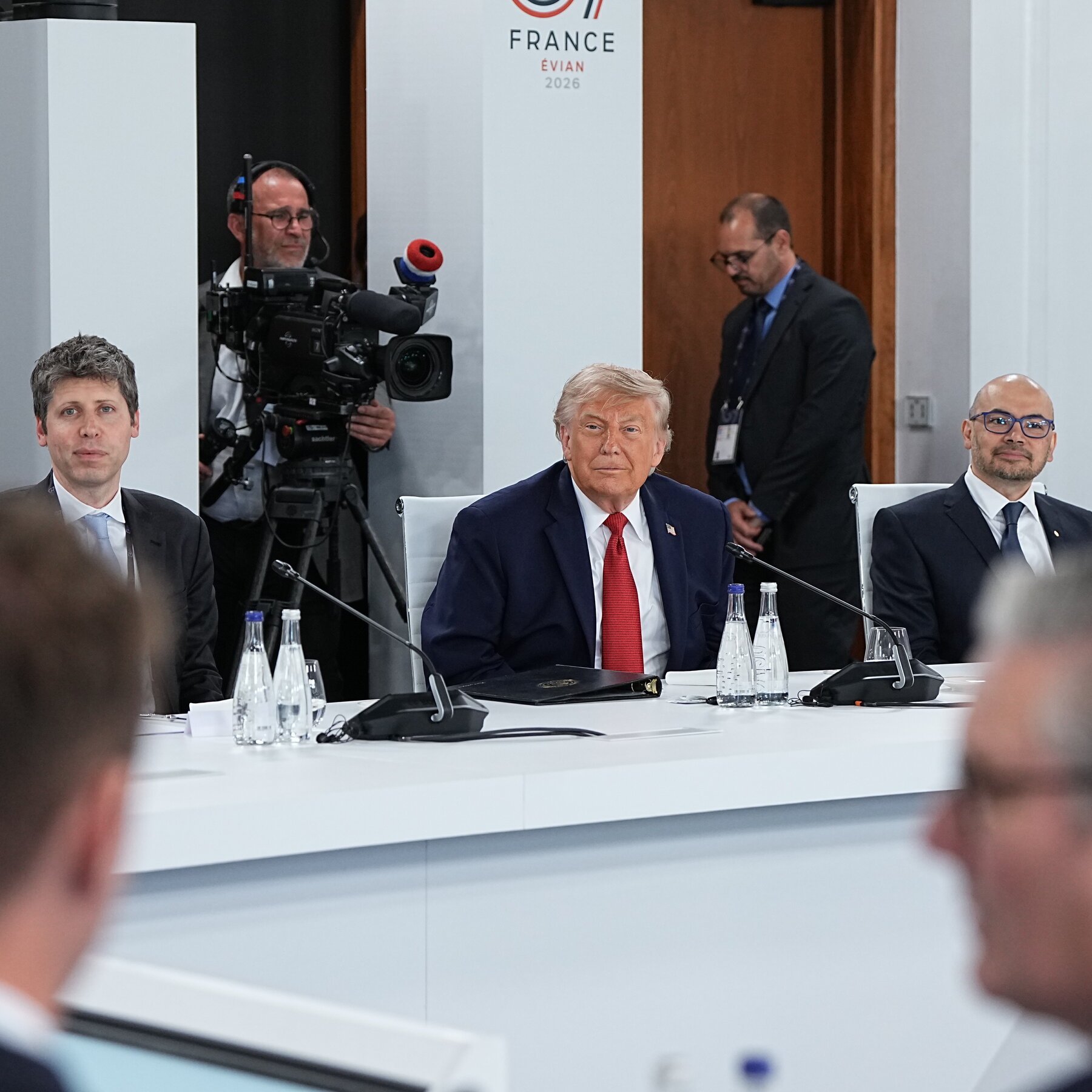 Three men in suits sit at a white conference table with water bottles and microphones, with a camera operator and a "G7 FRANCE ÉVIAN 2026" banner visible behind them.