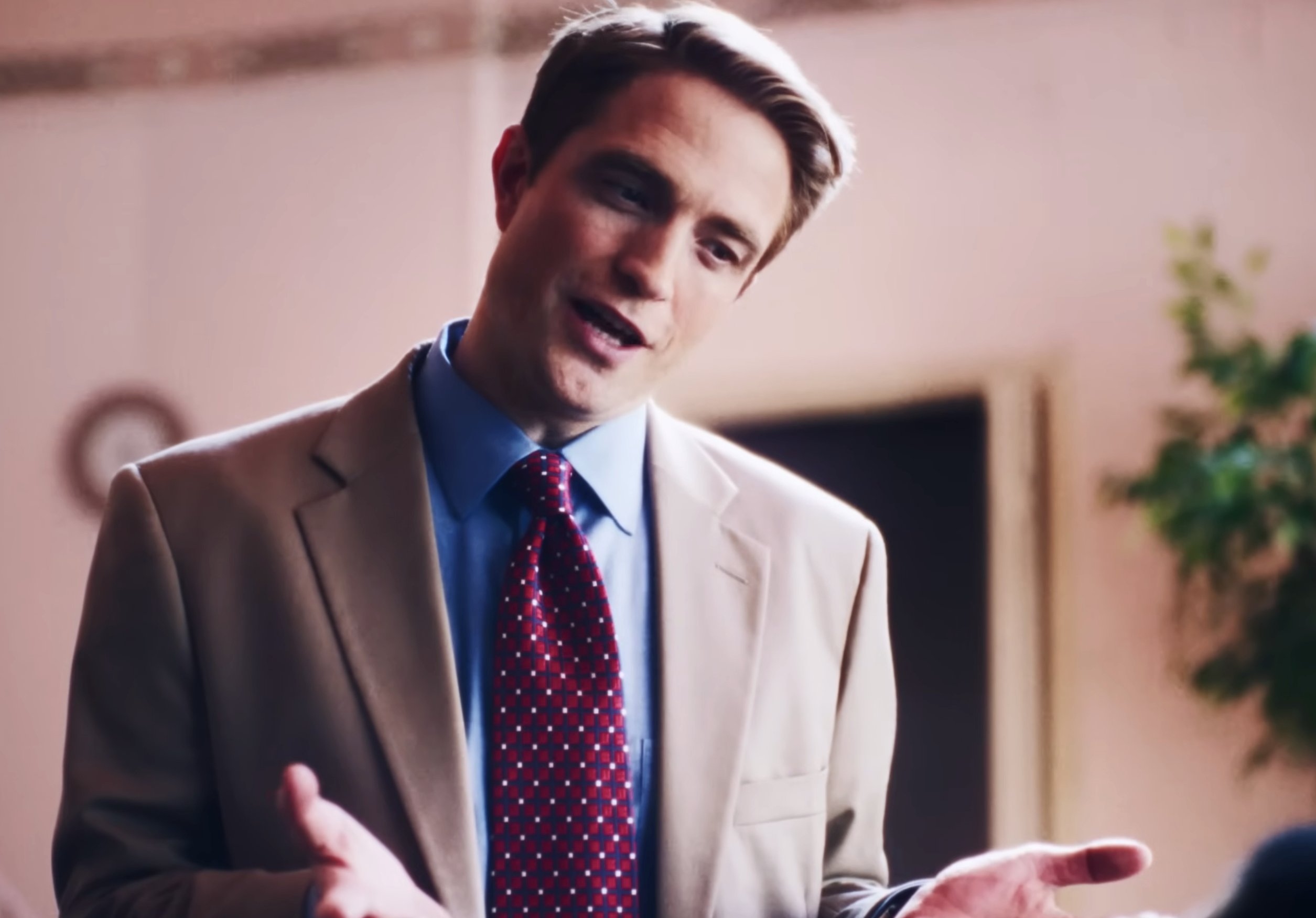A man in a beige suit, blue shirt, and patterned red tie speaks while gesturing with his hands indoors near a potted plant.