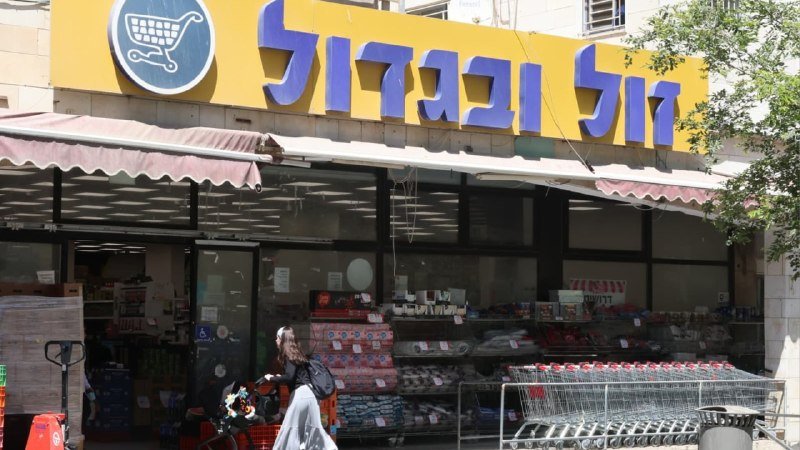 A storefront with a yellow sign displaying a shopping cart logo and Hebrew text features stacked merchandise, shopping carts, and a woman with a stroller outside.