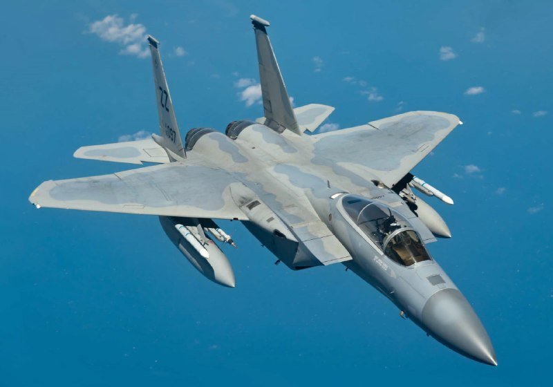 A gray fighter jet with twin tails, marked "SJ" and "350," banks sharply in flight over deep blue water, carrying external fuel tanks and missiles.