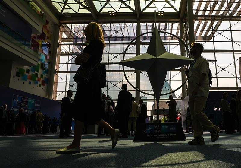 Attendees walk past a large compass-rose sculpture displayed inside a glass-walled venue, with signage reading "NATO Summit Ottawa Defence Industry Forum" visible at the base.