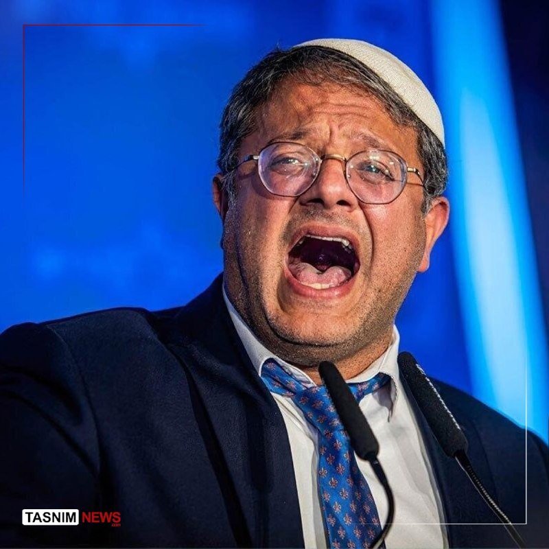 A bespectacled man wearing a white skullcap, dark suit, and patterned blue tie speaks animatedly into two microphones against a blue-lit backdrop, with a "Tasnim News" logo visible.