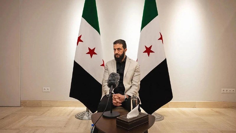 A bearded man in a light blazer sits at a small table speaking into a microphone, flanked by two flags bearing red stars on green, white, and black stripes.