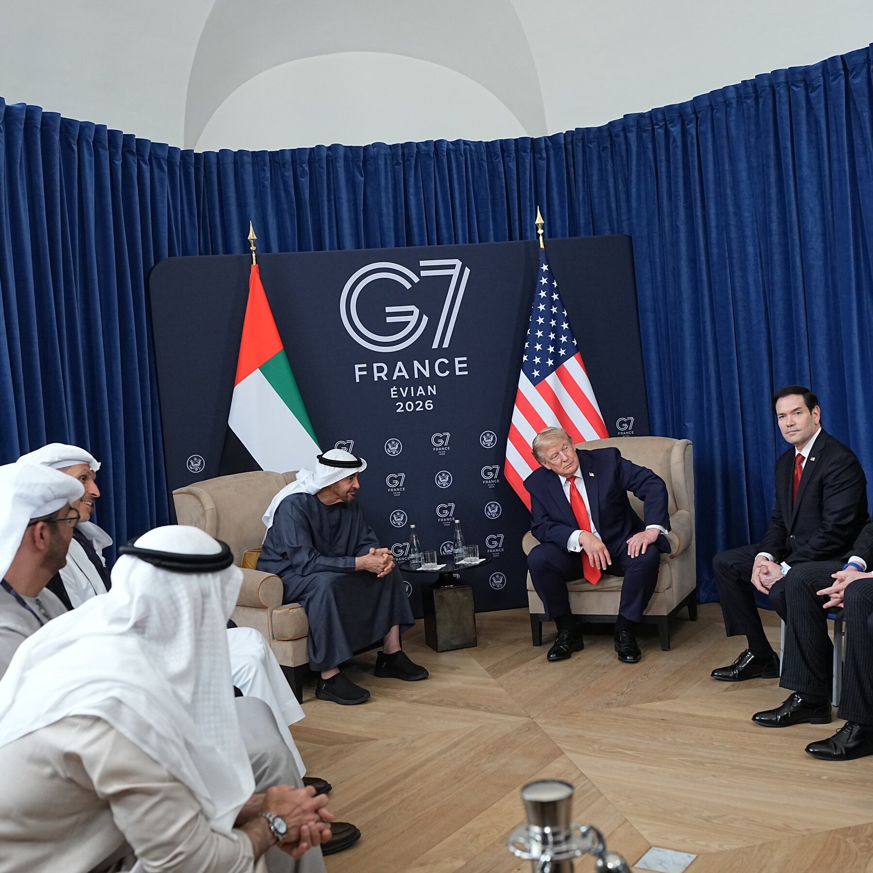 A bearded man in a black shirt speaks in front of blurred blue, yellow, and red-white flags.