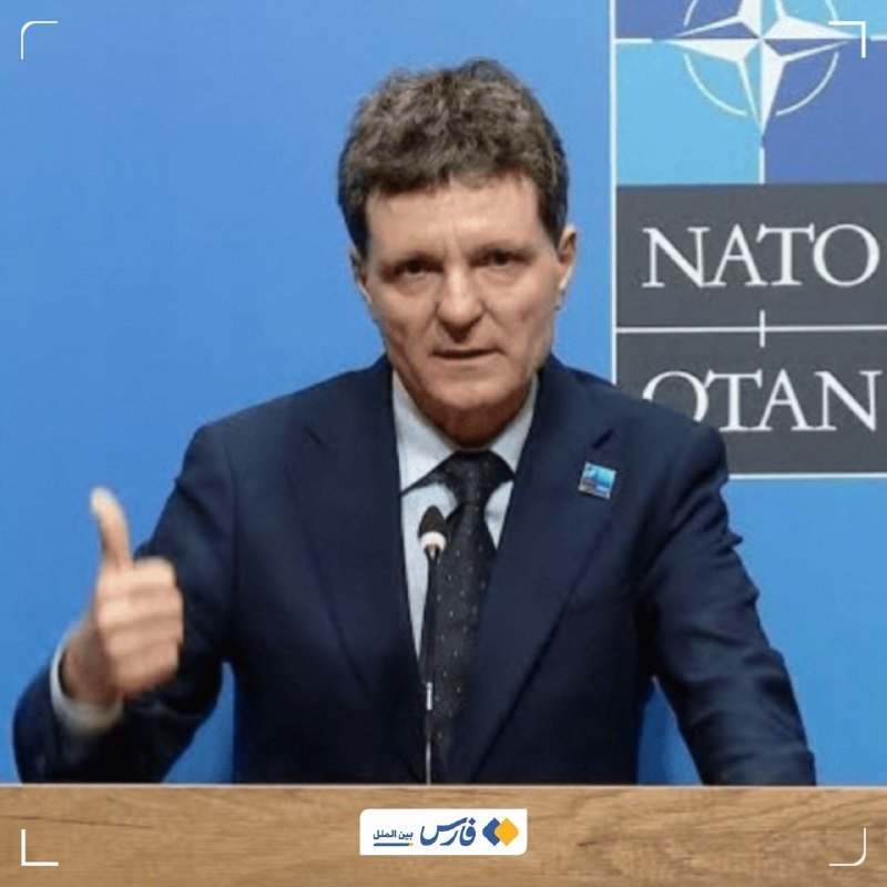 A man in a dark suit speaks at a podium, gesturing with his right hand, in front of a blue backdrop displaying the NATO logo.